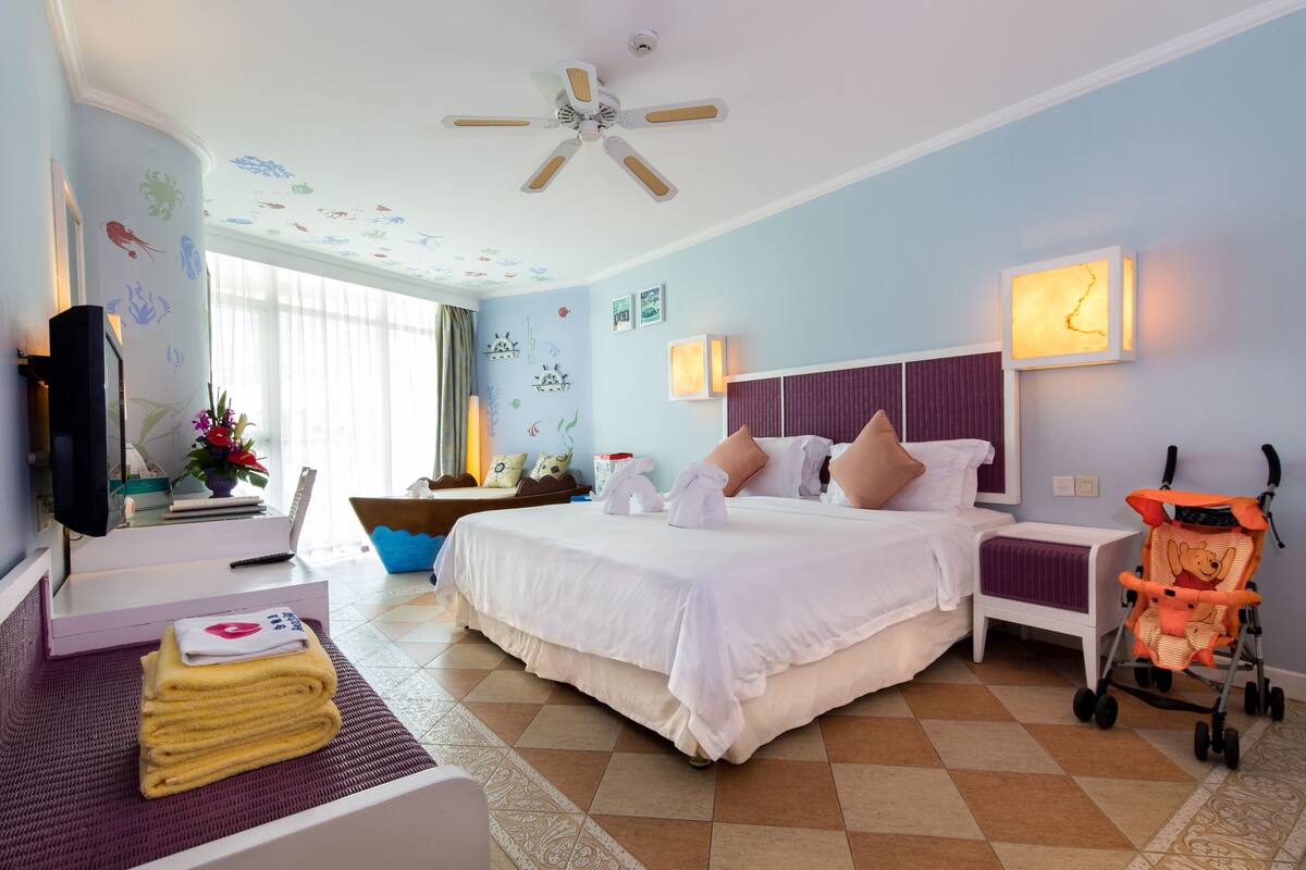 Habitación de hotel con cama, televisión, escritorio, silla, ventana y ventilador de techo.