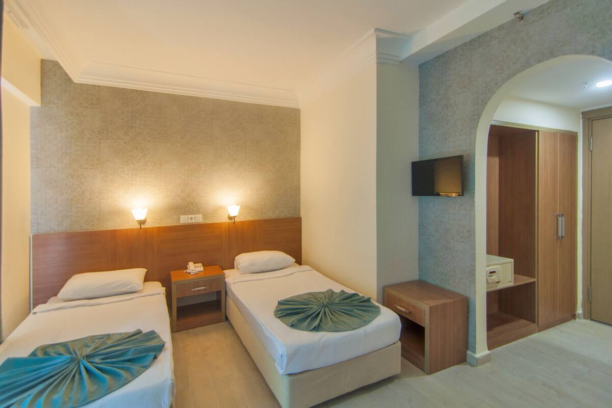 A hotel room with two beds, bedside lamps, a TV mounted on the wall, a wooden wardrobe, and a small bedside table.