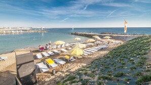 Een strand met rijen ligbedden en parasols, een rotsachtige kustlijn en helder uitzicht op de zee.