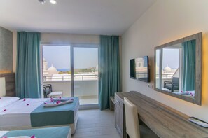 A hotel room with two beds, a desk, a TV, and a balcony with a view of buildings. - Kolibri Hotel (Alanya)