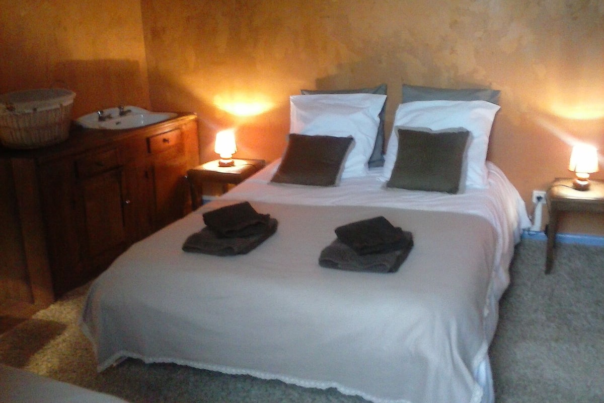 Uma cama bem arrumada, com duas toalhas, uma cômoda de madeira e uma mesa de cabeceira com abajur.