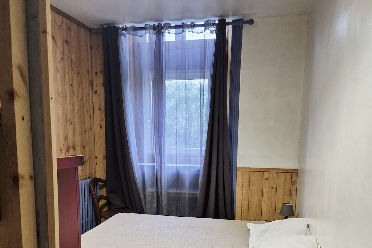 Quarto com cama, janela com cortinas, abajur sobre uma mesa de cabeceira e painéis de madeira nas paredes.