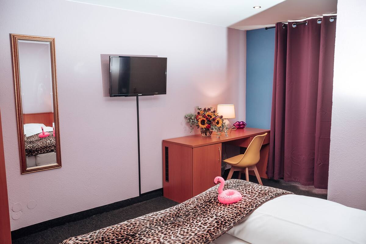 A hotel room with a bed, a desk, a chair, a TV, a mirror, and a window with curtains.