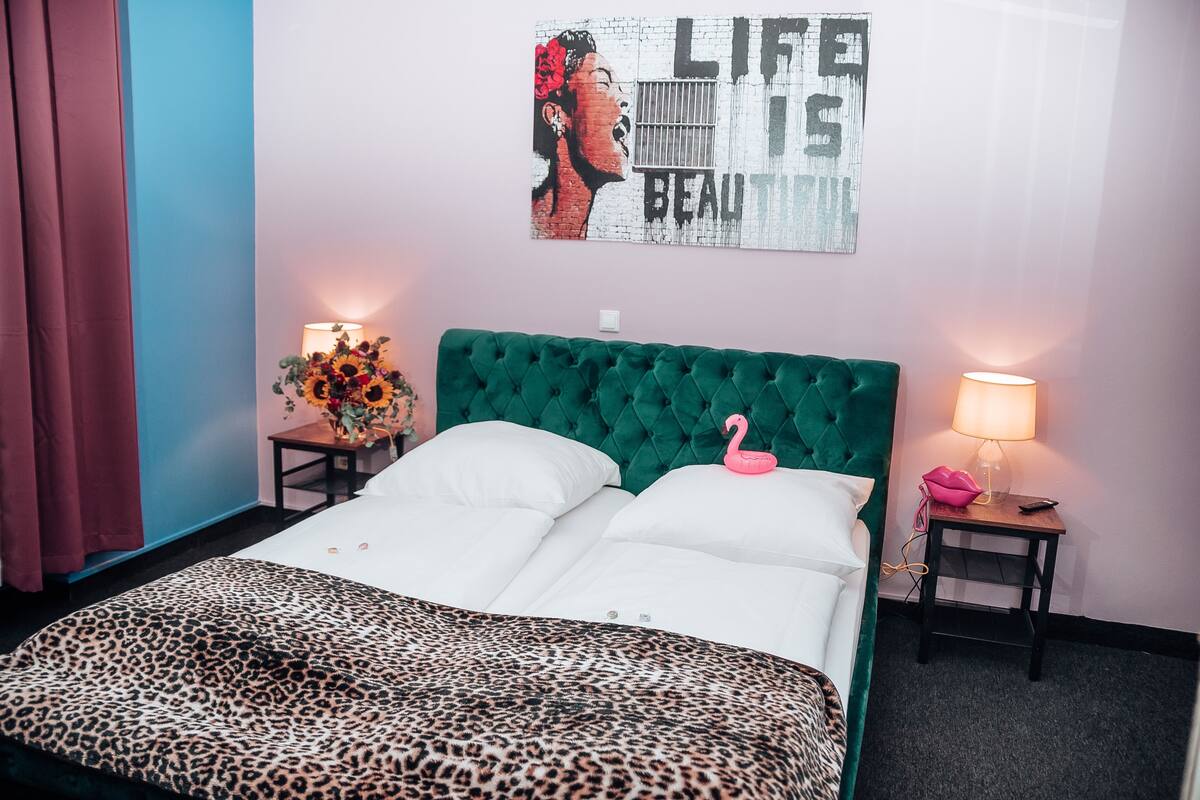 A bedroom with a green tufted headboard, a leopard print bedspread, a wall art piece, and two bedside tables with lamps and a vase of sunflowers.