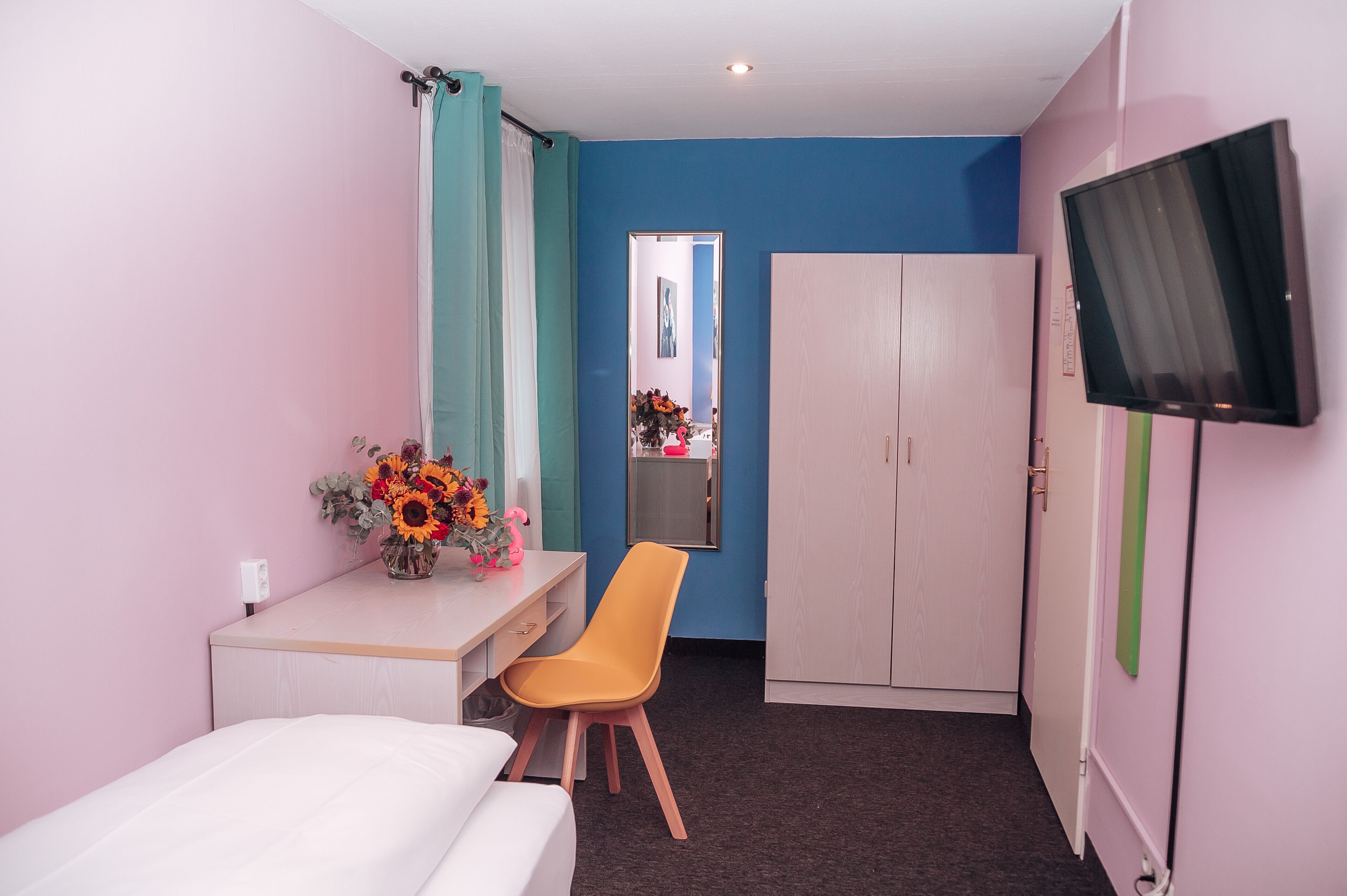 A hotel room with a bed, a desk with a chair, a wardrobe, a TV, and a mirror.