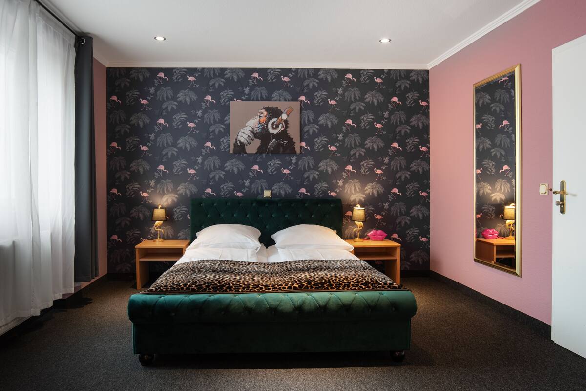 A hotel room with a large bed, two bedside tables with lamps, a mirror, and a wall with a tropical wallpaper design.