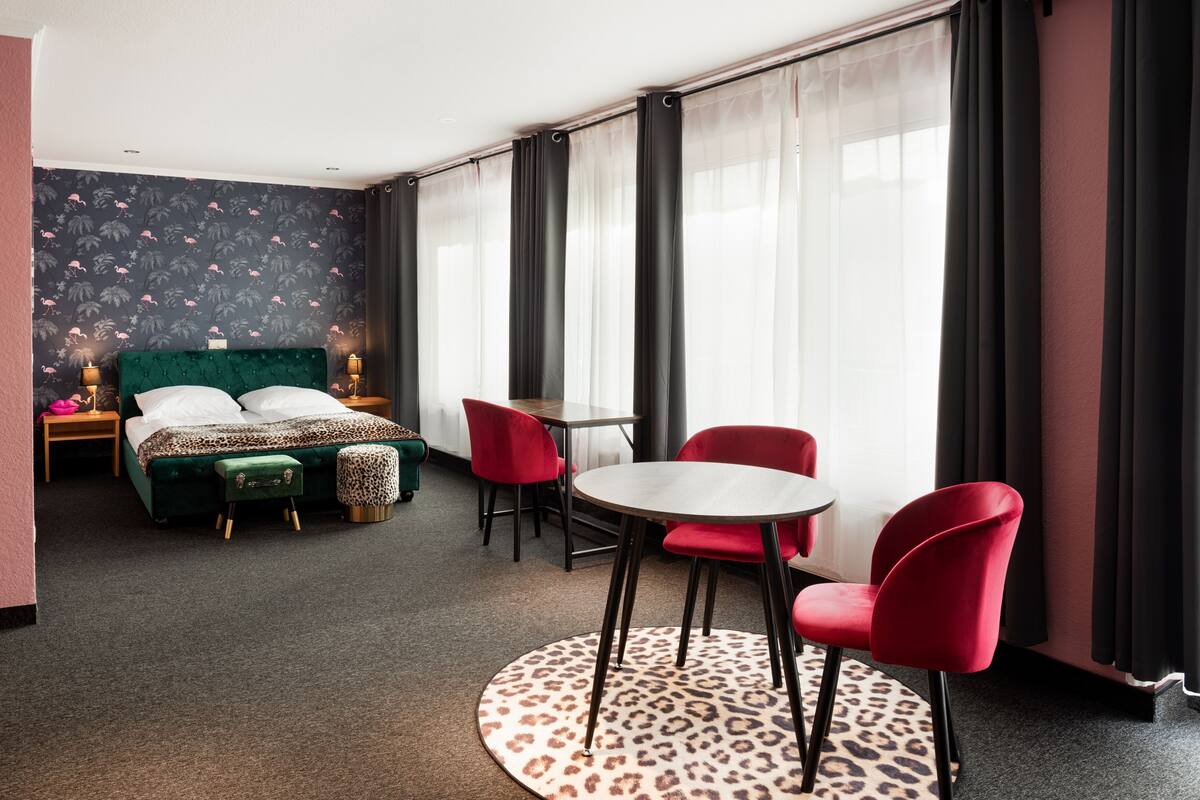 A hotel room with a bed, a round table, red chairs, a leopard print rug, and sheer curtains.