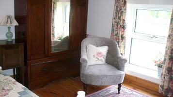 Una habitación con un aparador de madera, un sillón gris con un cojín floral, un cabecero de madera y una ventana con cortinas estampadas de flores.