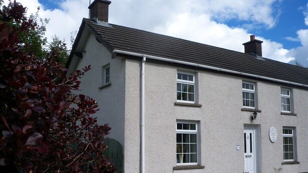 A two-story house with a white exterior, a grey roof, and a green door. There are two windows on the ground floor and one on the upper floor. - Lisnafillan Lodge (Ballymena)