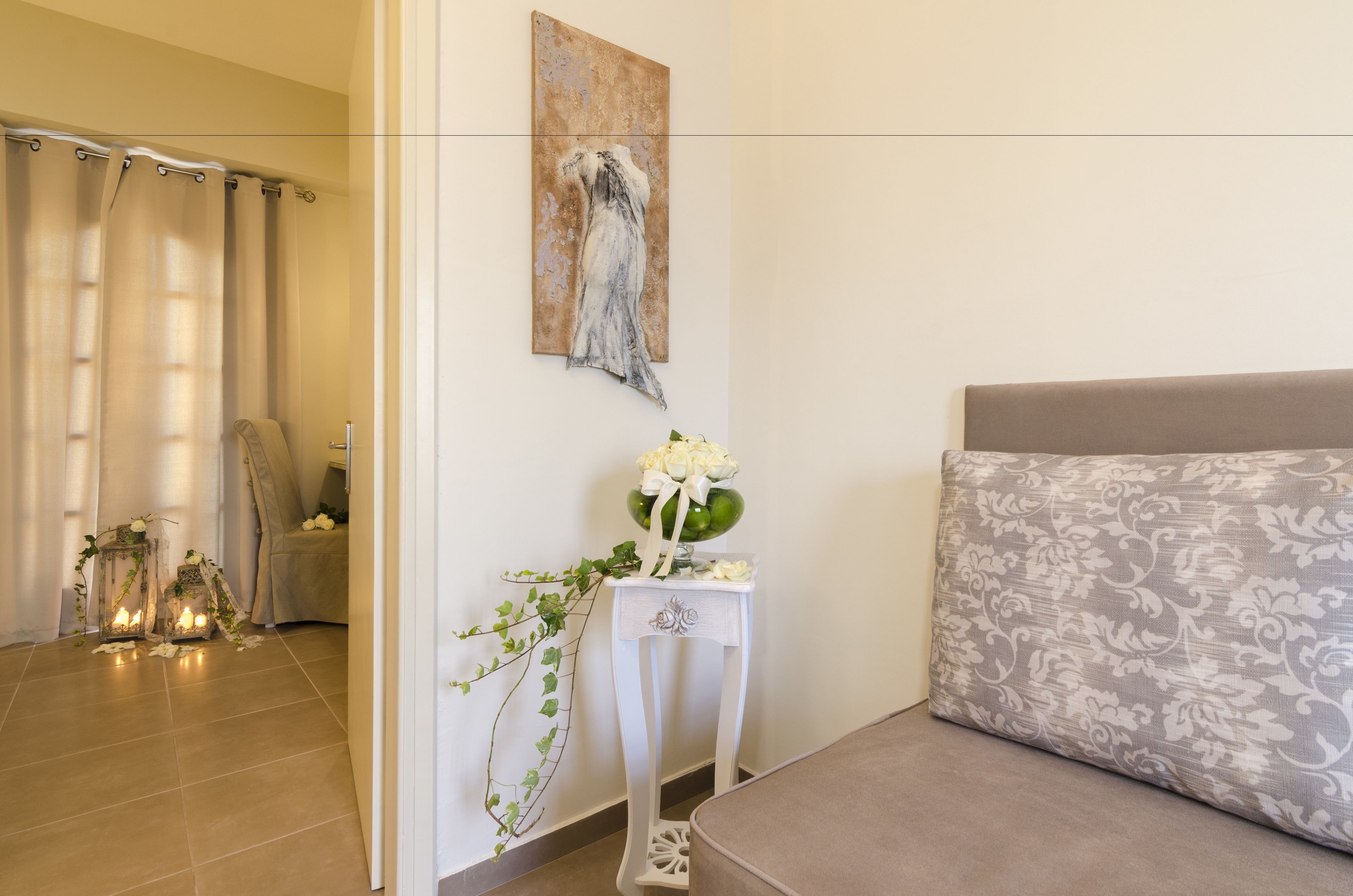 Une pièce avec un canapé beige, un élément décoratif mural, une petite table blanche ornée d’un vase de fleurs et un rideau.