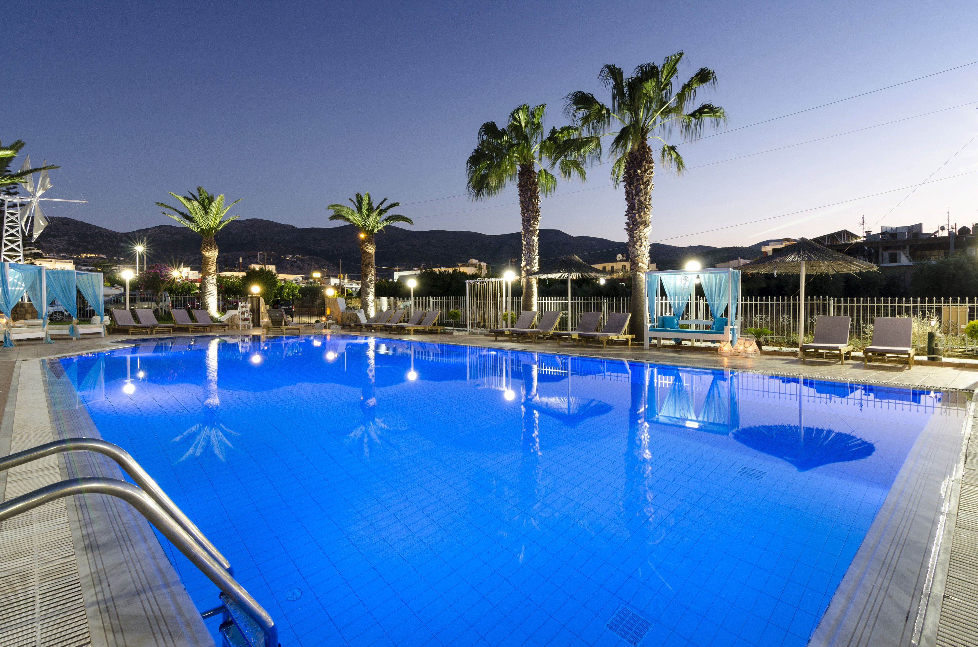 Piscina con sillas de descanso y palmeras, rodeada por una valla e iluminación.
