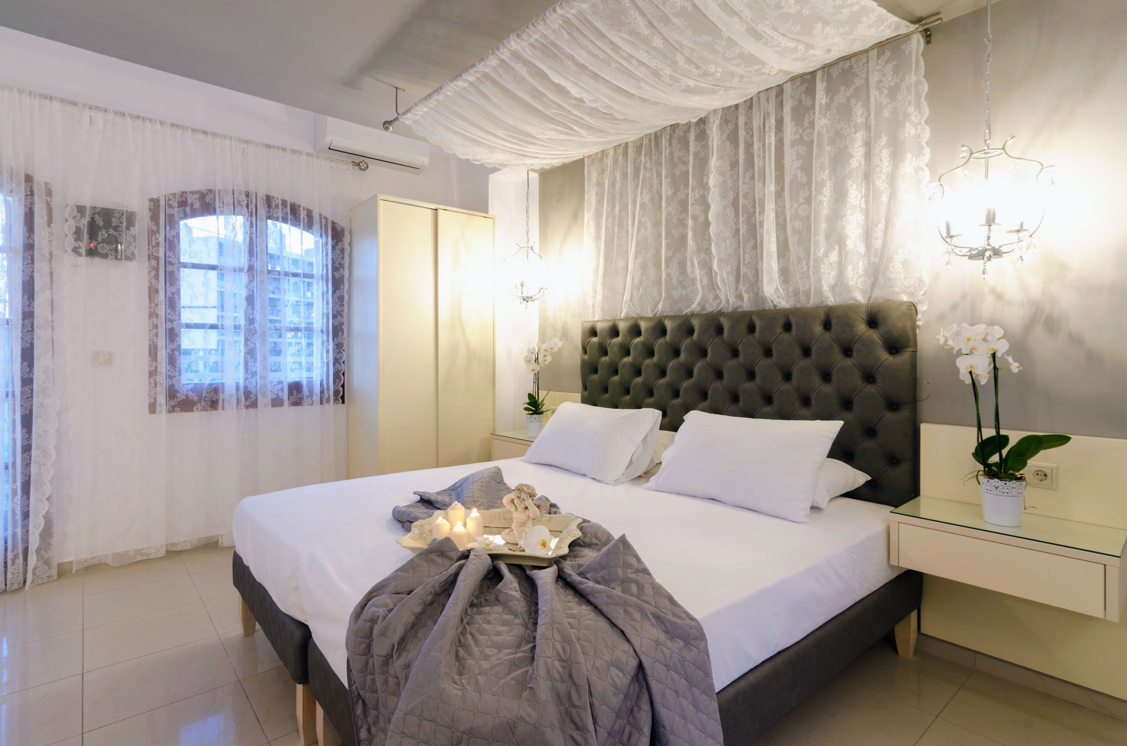 A neatly made bed with a grey quilt, a bedside table with a vase of flowers, and a chandelier.