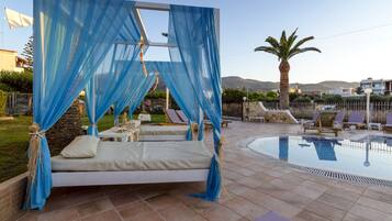 Zona de piscina con cama y sillones de descanso, rodeada por una piscina y palmeras.