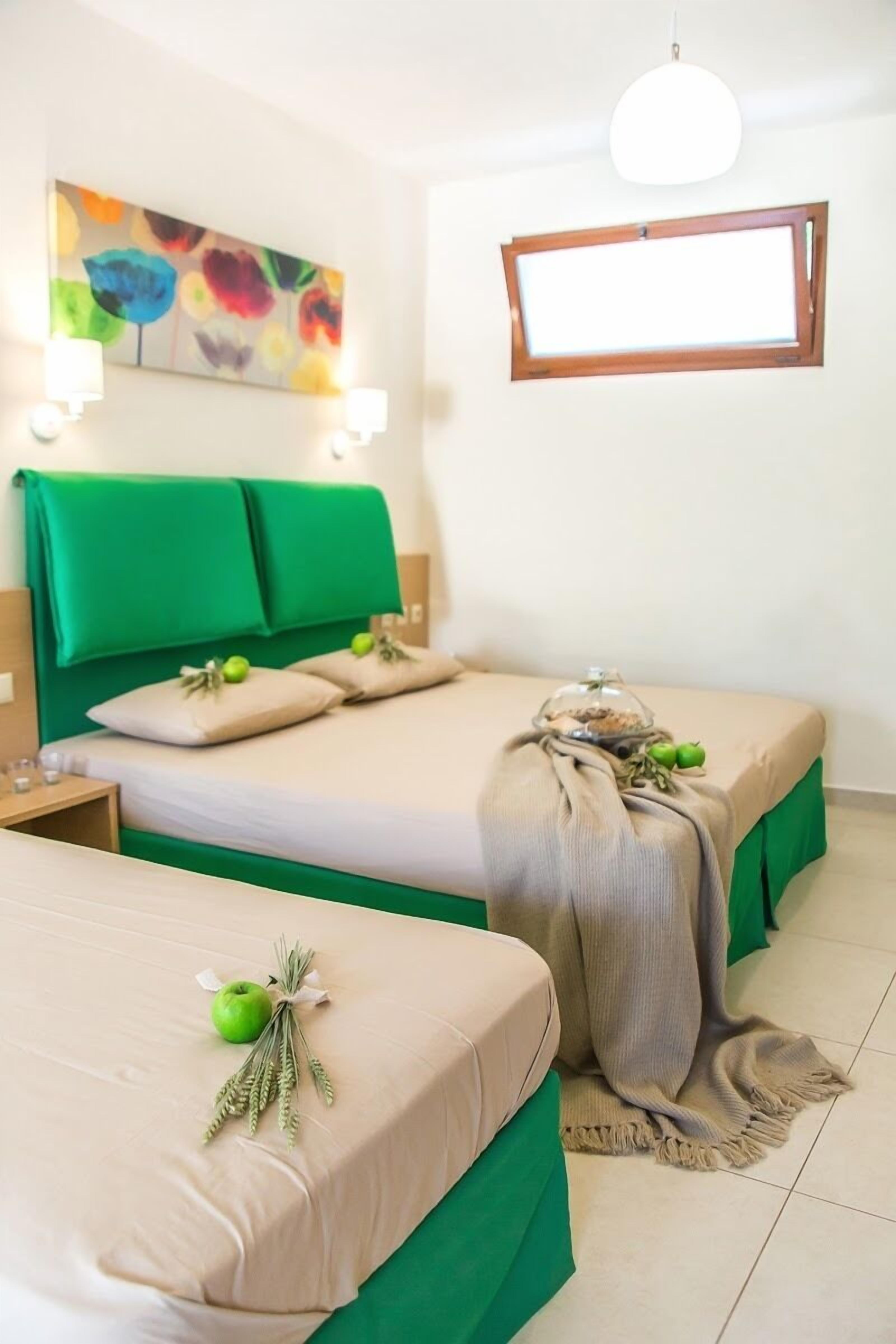A hotel room with two beds, green headboards, a colorful floral painting, a window, and a small table with a lamp.