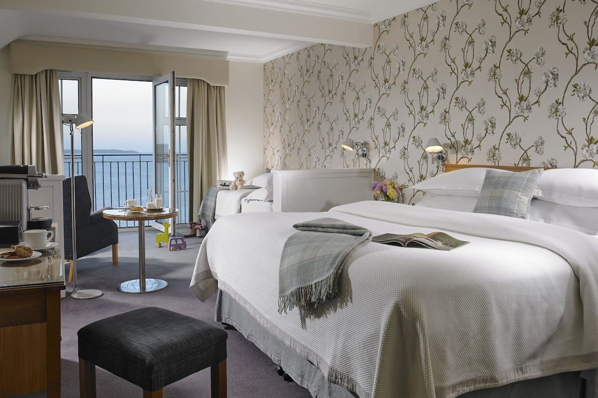 A hotel room with a bed, a desk, a chair, a small table, and a view of the sea through a balcony.