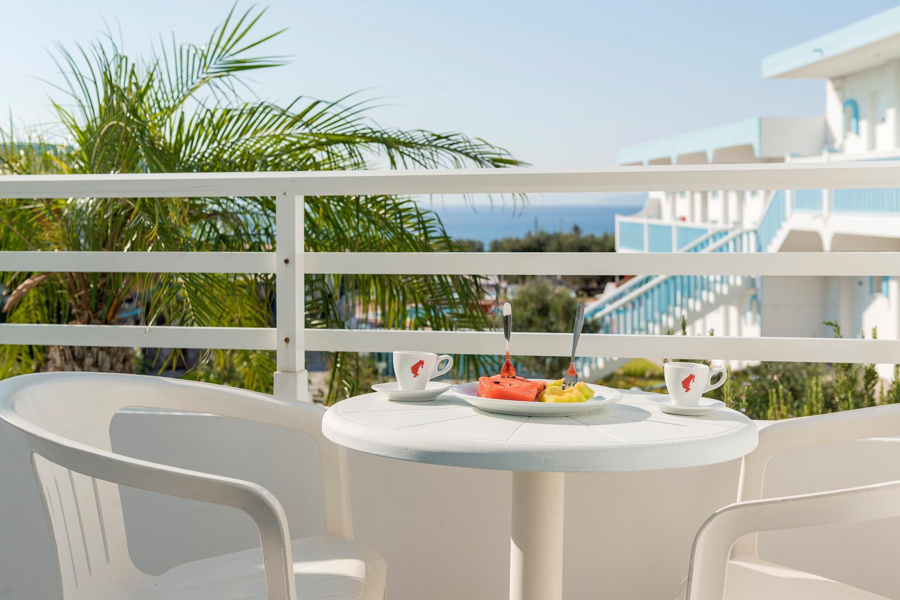 A white table with two chairs, a plate with food, and two cups on a balcony overlooking a blue building and palm trees.
