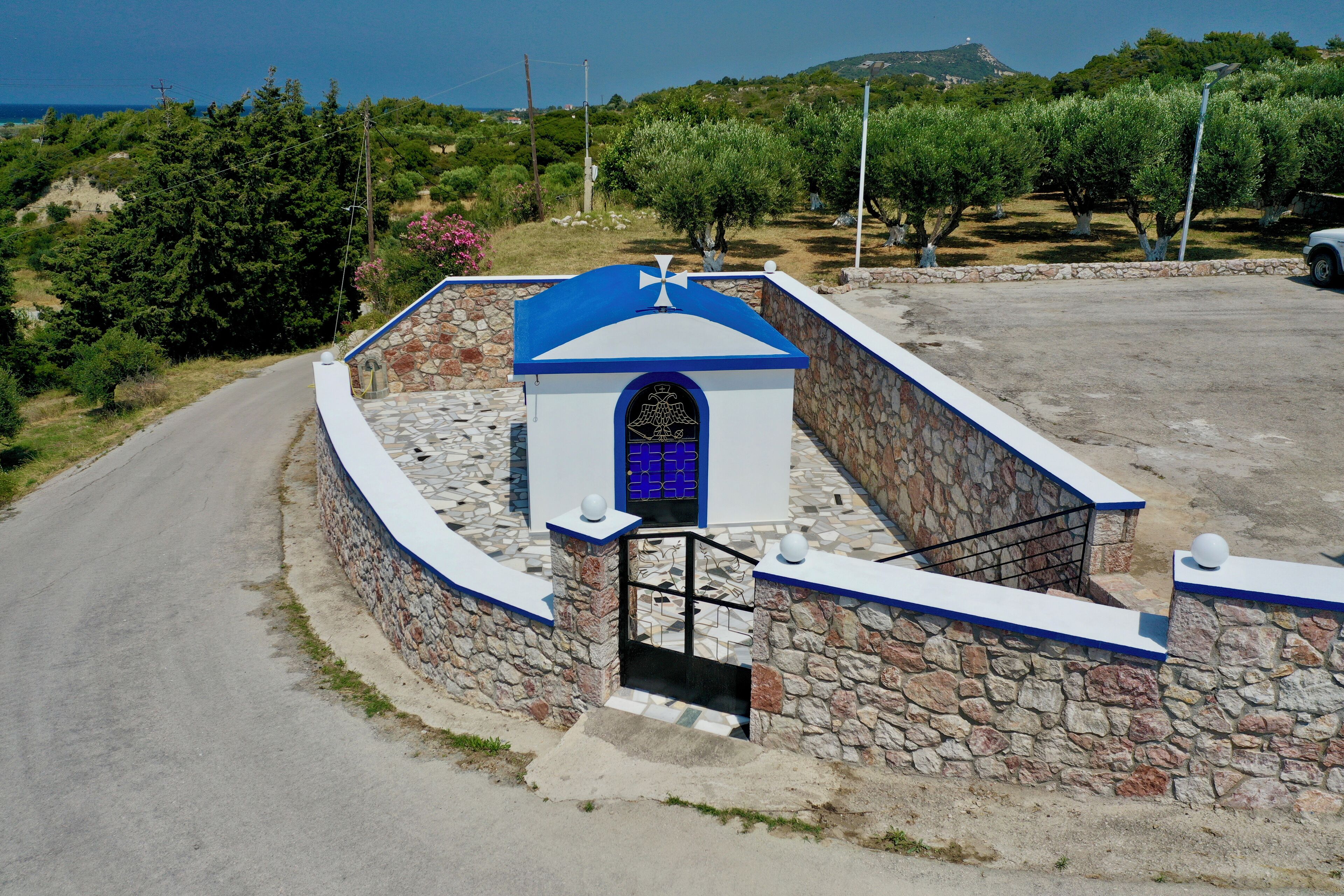 Un edificio de paredes de piedra con techo azul y una verja, rodeado de follaje y una carretera.
