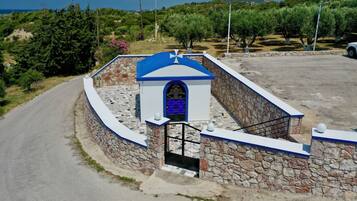 A stone-walled building with a blue roof and a gate, surrounded by greenery and a road.