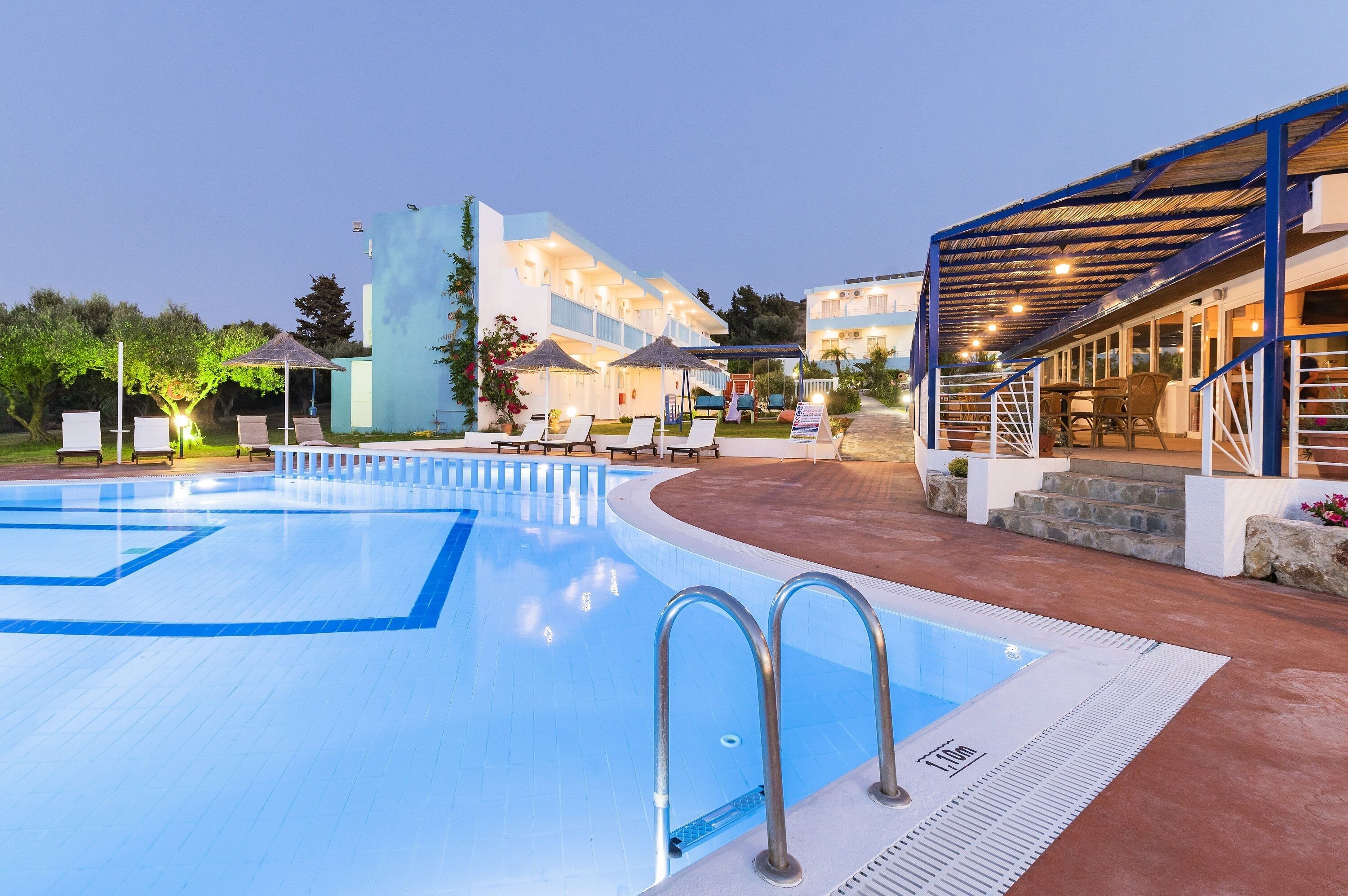 En swimmingpool med solstolar och parasoller, ett avskärmat sittområde och en byggnad med flera balkonger.