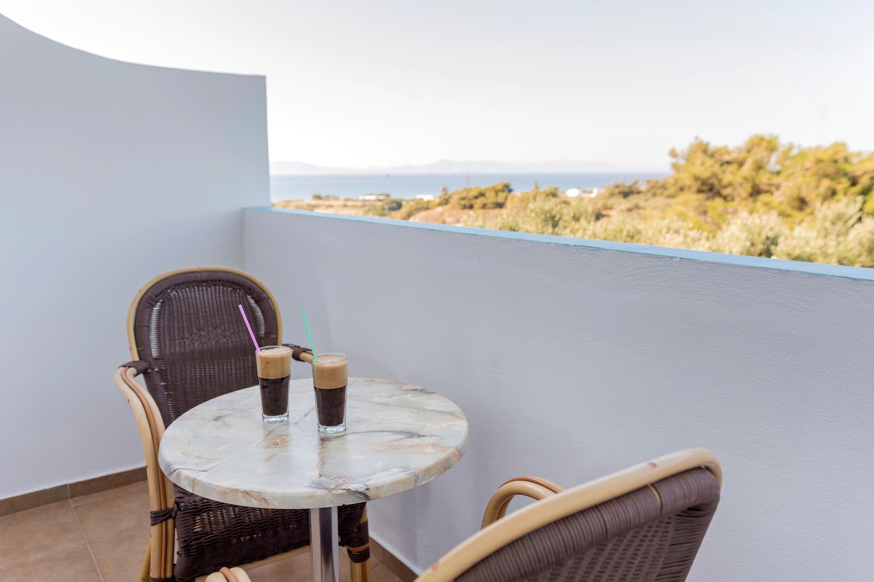 A balcony with a round table and wicker chairs, a glass of iced coffee, and a view of the landscape.