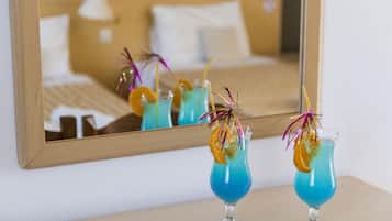 Two cocktails with garnishes on a table in front of a bed and a mirror reflecting the room.