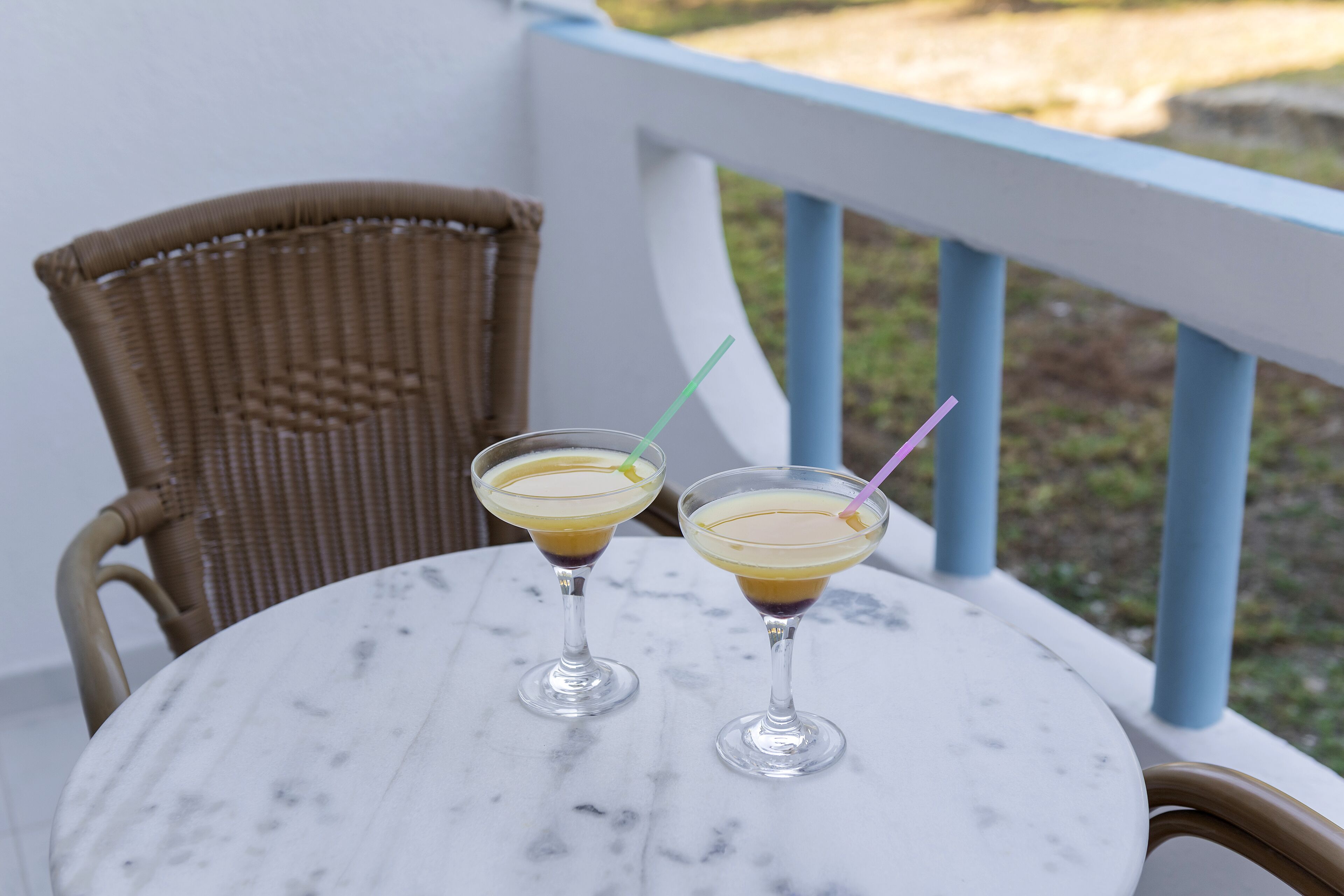 Two drinks on a marble table with a wicker chair beside it, on a balcony with a railing.