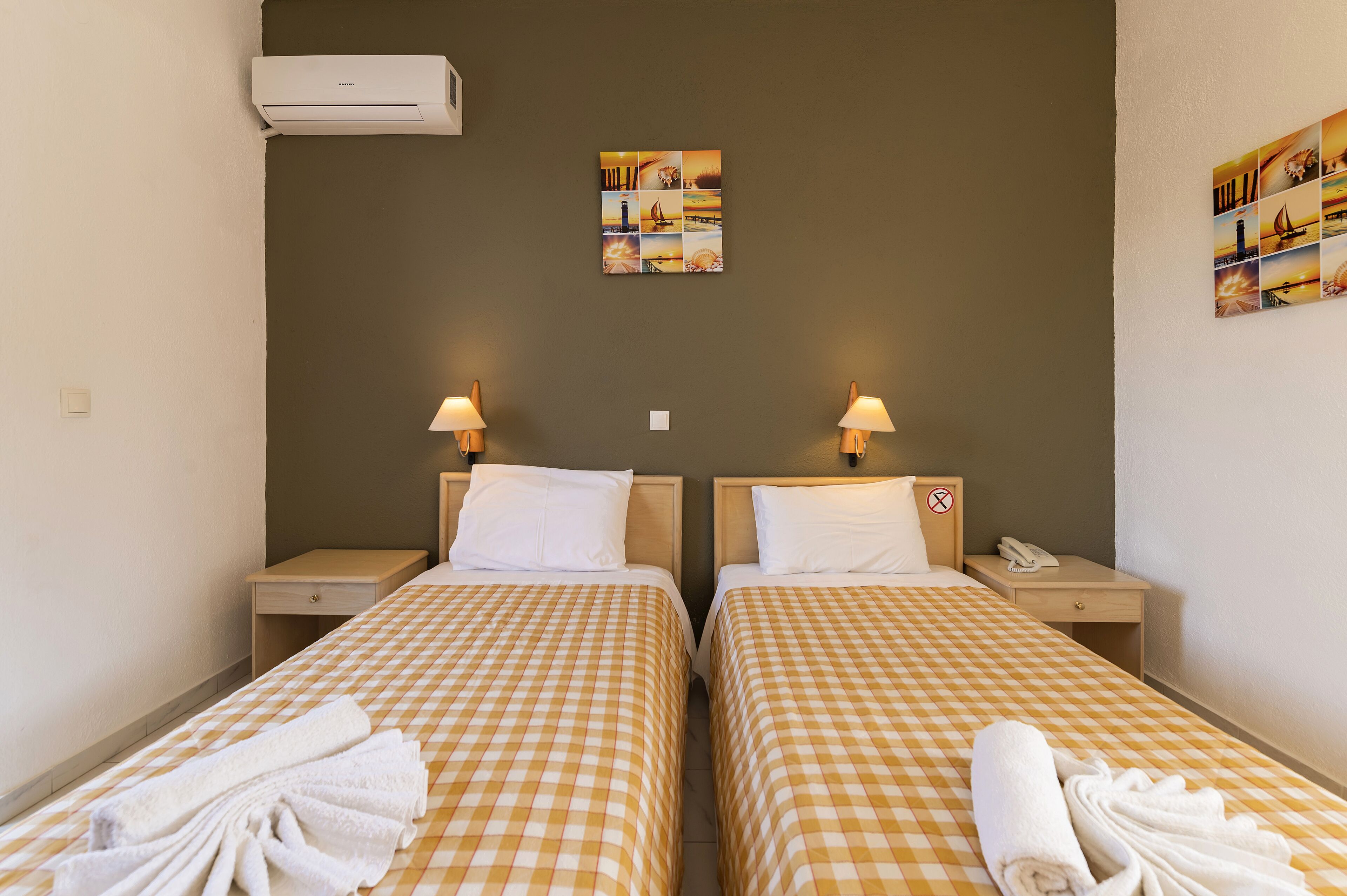 A hotel room with two beds, checkered bedding, a wall-mounted air conditioner, bedside lamps, a small table with a phone, and a wall art piece.