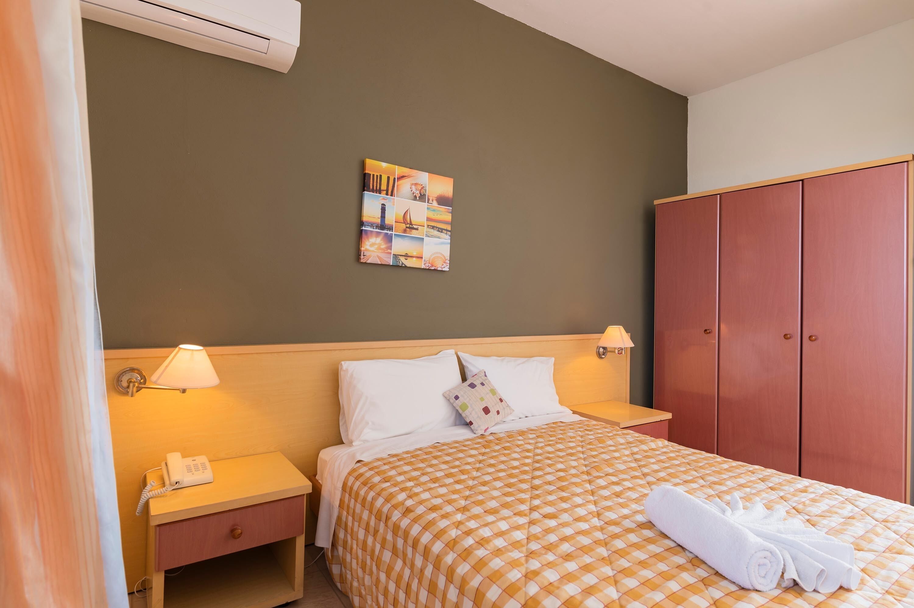 A hotel room with a checkered bedspread, a bedside lamp, a phone on the nightstand, a painting on the wall, and a wardrobe.