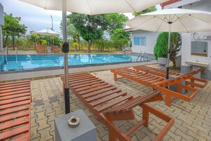 Piscina con sillones de descanso y sombrillas, rodeada de un área pavimentada con árboles y un edificio con un letrero.