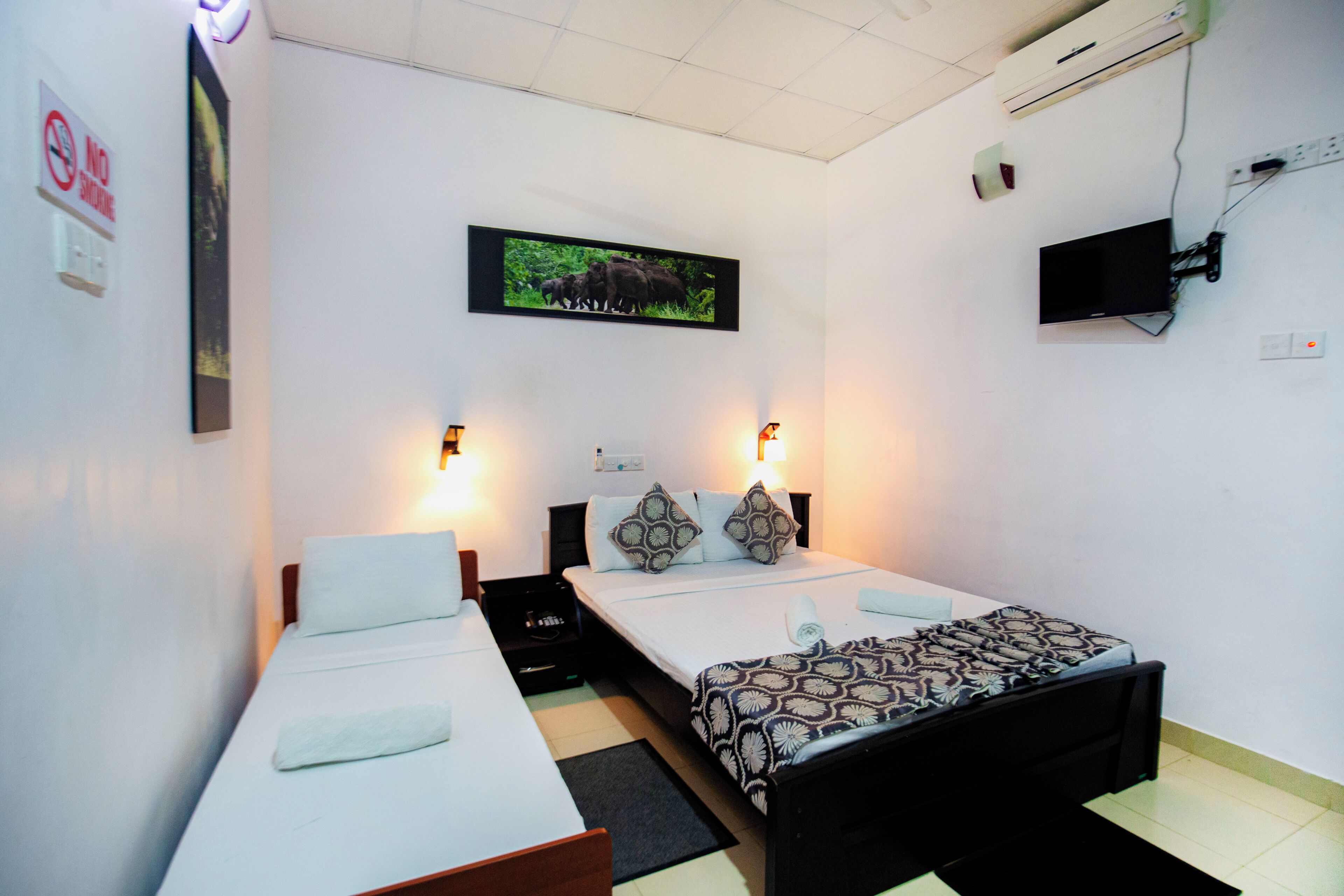 A hotel room with two single beds, a flat-screen TV, a mounted air conditioner, and a framed picture of elephants on the wall.