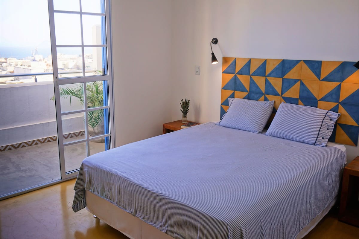 Een slaapkamer met een bed, nachtkastje, lamp en uitzicht op de stad door een raam.