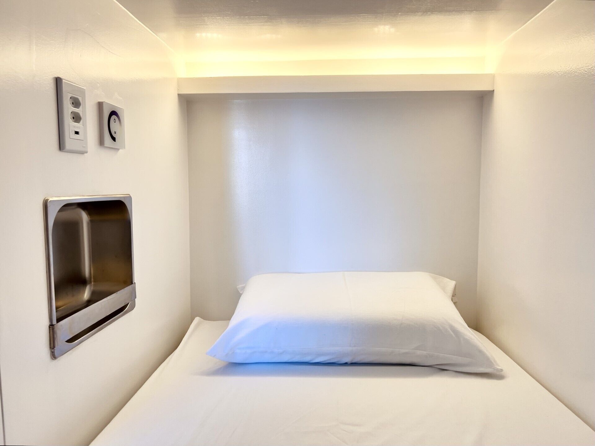 A small, single-bed room with a white mattress and pillow, a built-in sink, and a wall-mounted control panel.
