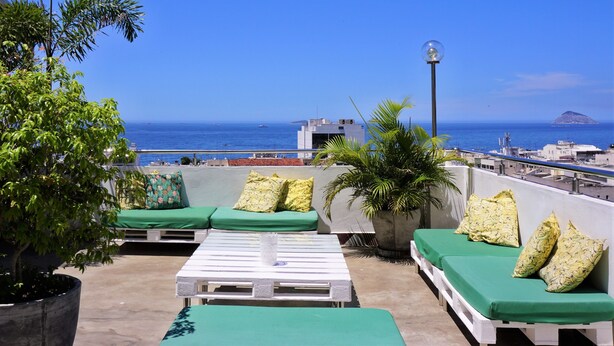 Une terrasse sur le toit, dotée de sièges confortables avec des coussins verts, d’une table blanche et d’une plante en pot.