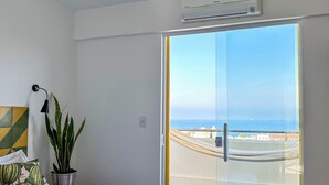 Une chambre à coucher avec un lit, une plante en pot, une lampe fixée au mur et une vue sur la mer, visible à travers une porte coulissante.