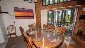A dining area with a wooden table and chairs, a painting of a sunset, and a view of trees through large windows. - 1360 Knoll Lane 3 Bedroom Home by RedAwning (South Lake Tahoe)