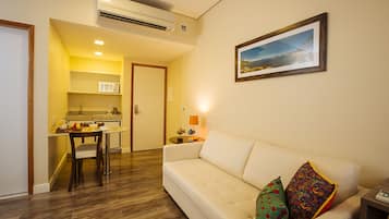 Un salón con un sofá blanco, almohadones coloridos, un cuadro en la pared y una cocina al fondo.