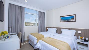 Quarto de hotel com duas camas, uma mesa de trabalho com computador, luminária e janela com cortinas.