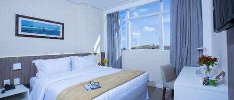 A hotel room with a bed, a desk, a chair, a window with curtains, a picture on the wall, and a vase with flowers.
