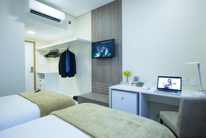 Quarto de hotel com duas camas, uma escrivaninha com um laptop, uma televisão e um minibar.