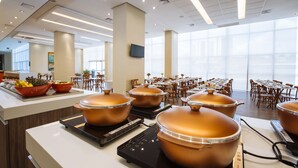 Un comedor moderno con varias mesas y sillas, ollas de cobre grandes sobre cocinas por inducción, y un amplio ventanal que proporciona luz natural.