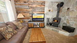 A cozy living room with a brown leather sofa, a wooden coffee table, a flat-screen TV, and a stone fireplace with a log fire. - 1510 Wildwood, 6 2 Bedroom Condo by RedAwning (South Lake Tahoe)