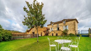 Une maison en pierre avec une clôture en bois, un arbre et des chaises en métal blanc dans la cour.