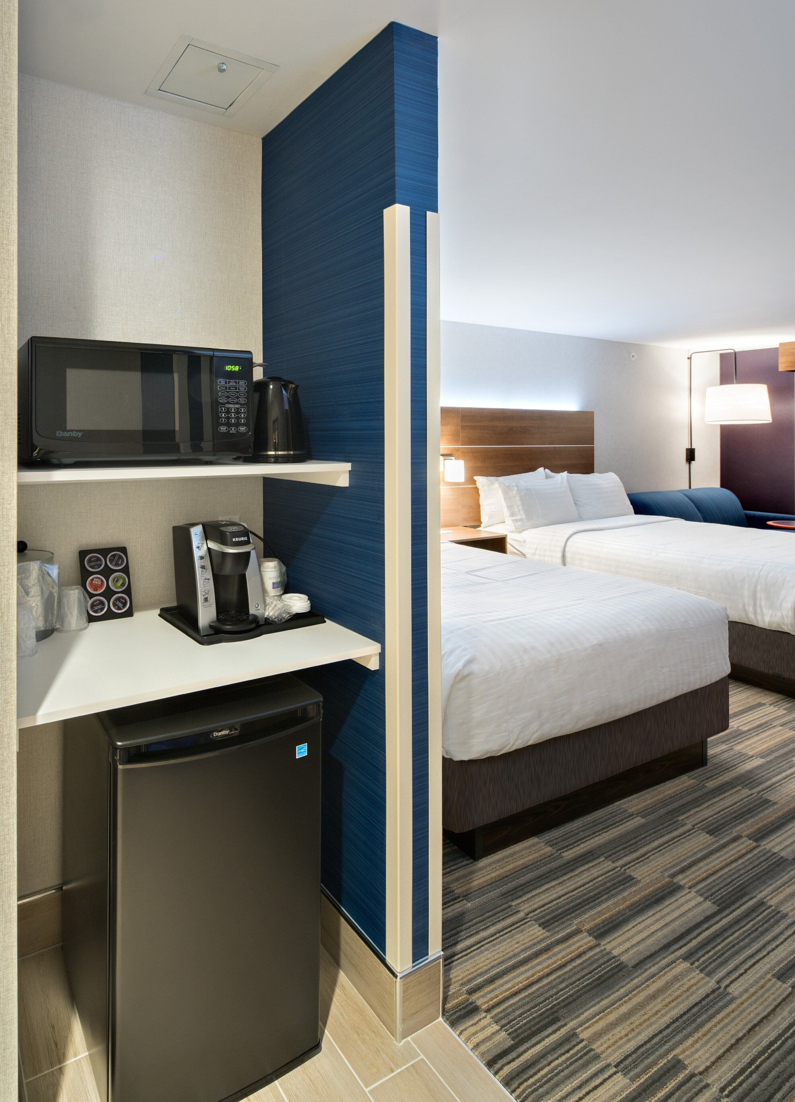 A hotel room with a microwave, coffee maker, and a compact refrigerator. There are two beds with white linens and a blue accent wall.