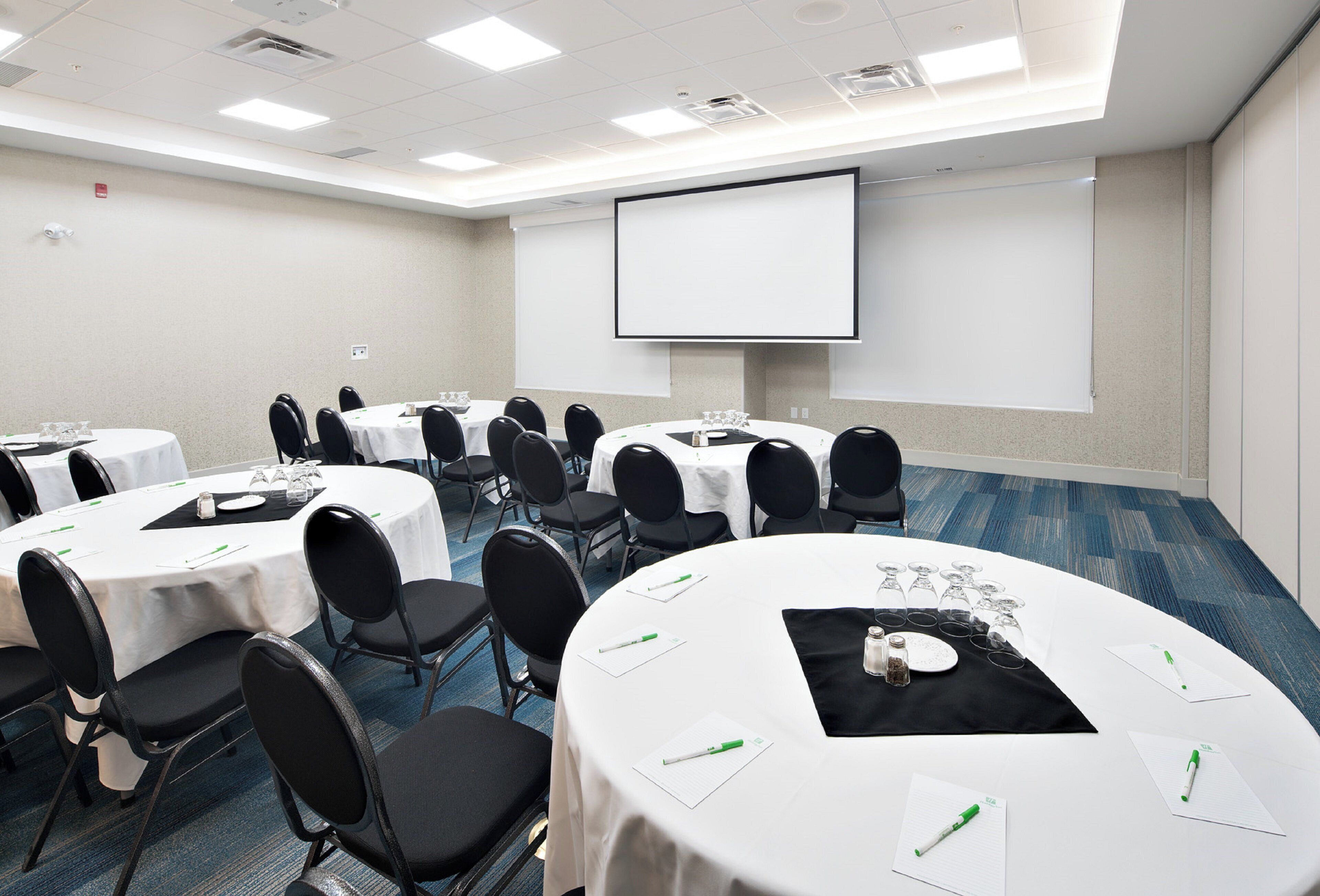 Un salón de conferencias con varias mesas, sillas, una pantalla de proyector y suelo alfombrado.