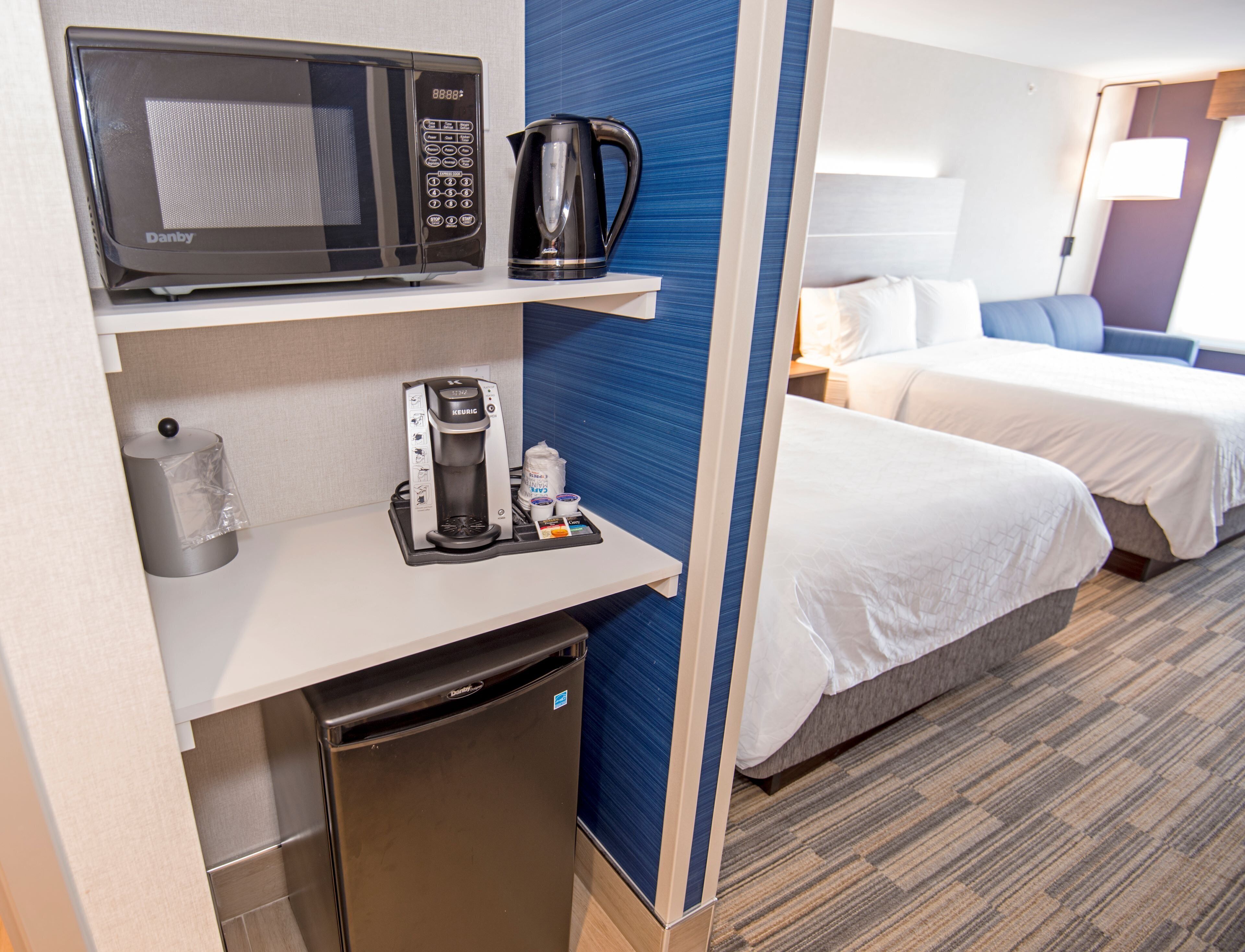 Habitación de hotel con microondas, cafetera y un refrigerador pequeño en la encimera. Se ve una cama con ropa blanca y una pared de acento azul.