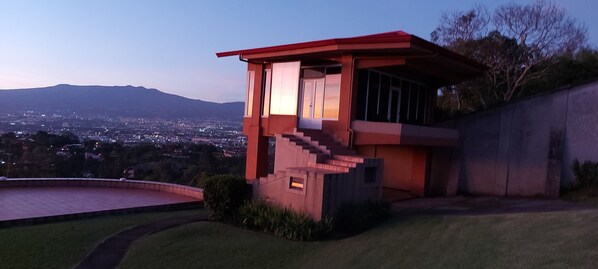 A modern house with a red roof, a staircase leading to the entrance, and a view of a cityscape in the background.