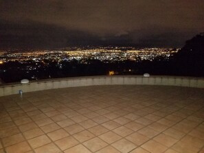 Vista panorámica de una ciudad de noche desde un punto elevado, con suelo de baldosas y barandilla.