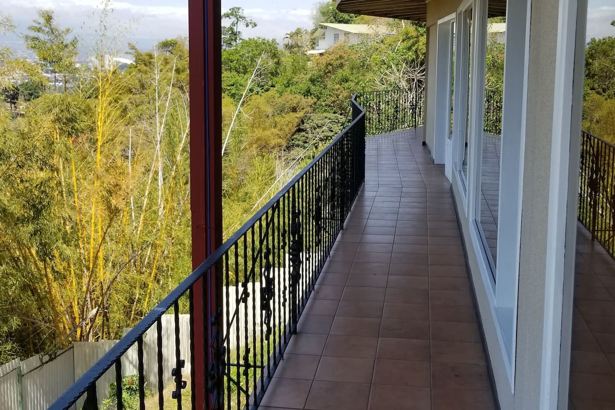 Ein überdachter Balkon mit Fliesenboden, Metallgeländer und Blick auf eine grüne Landschaft mit Bäumen und einem entfernten Gebäude.
