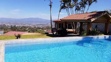 Relax by the serene pool surrounded by lush greenery and a distant cityscape.