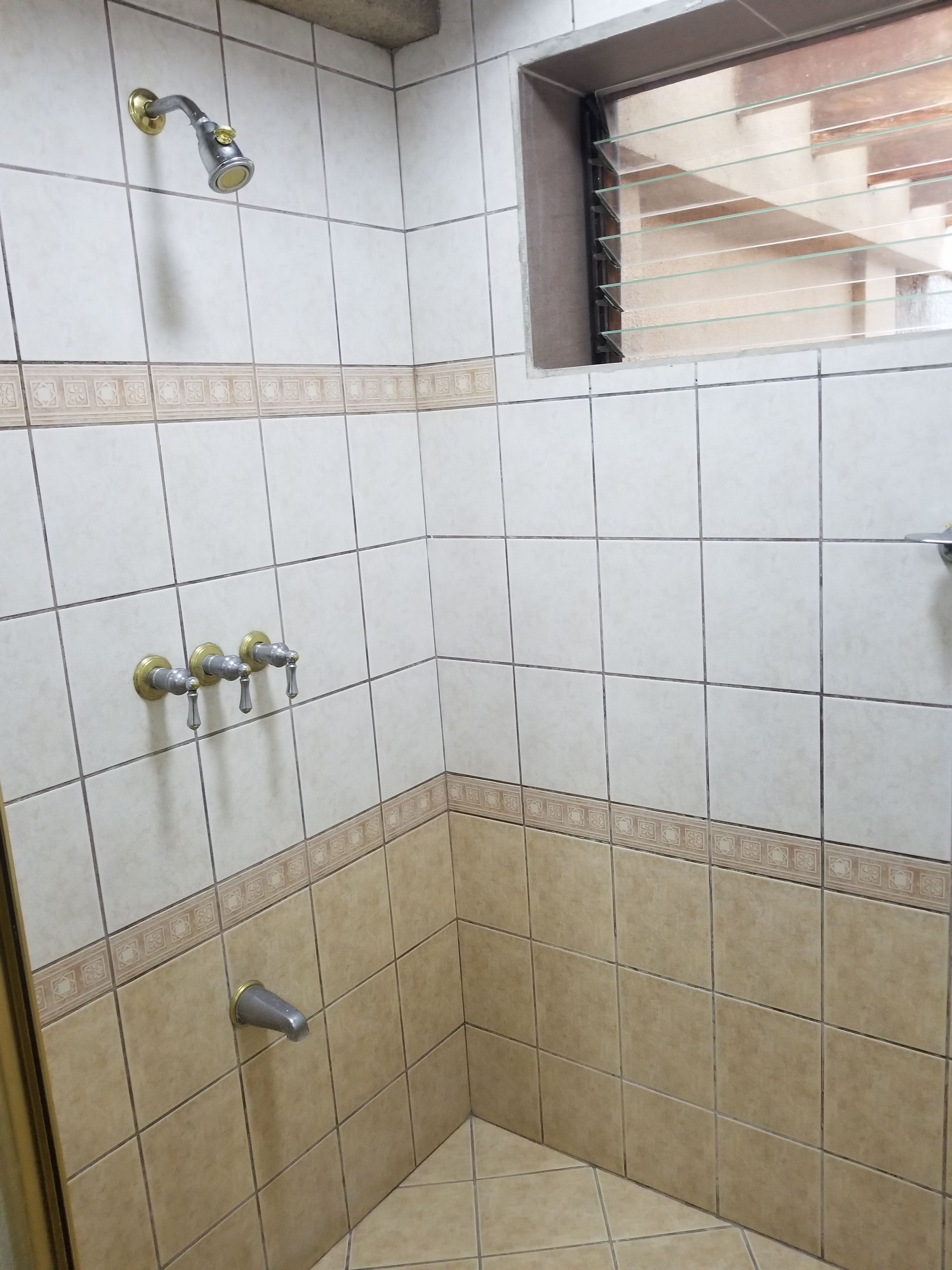 Una esquina de un baño con azulejos blancos y color crema, un cabezal de ducha y una ventana con celosía metálica.